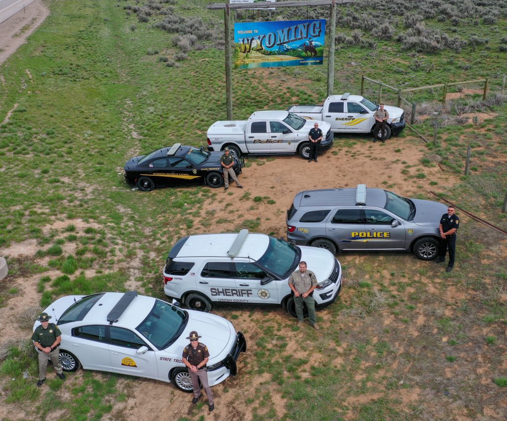 Officers from Wyoming Highway Patrol, Evanston Police Department, Uinta County WY Sheriff's Department, Vernal Police Department, Unitah County Sheriff's Department and Utah Highway Patrol stand by their vehicles with the Welcome to Wyoming sign in the background.