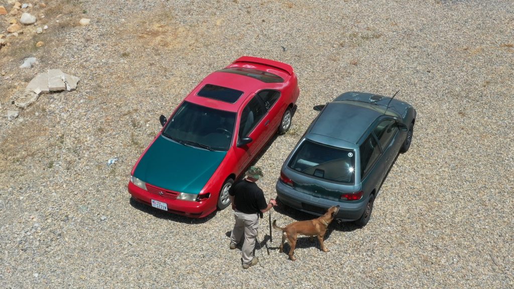 Aerial view of K9 Arros sniffing a vehicle as Trooper Colvin holds his leash.
