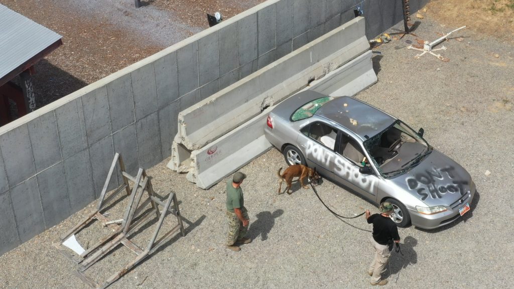 Aerial view of K9 Arros sniffing a vehicle as Trooper Colvin holds his leash as part of a bomb training scenario.