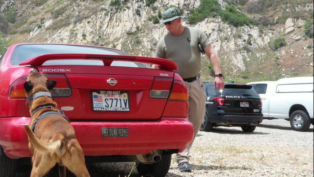 Trooper Elmer walks alongside a red car as K9 Rocco sniffs the trunk.