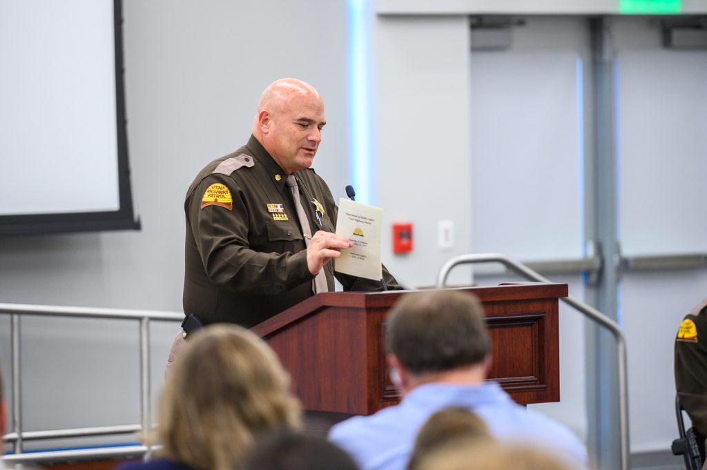 Major Winward stands at the podium delivering his remarks, holding a program that has the UHP values on the back of it.
