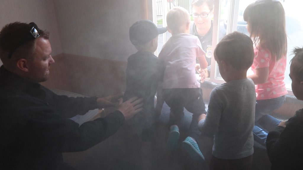 Children stand at the window of the Life Safety House Trailer as fake smoke fills the room and deputy fire marshal explains to them what to do. One deputy fire marshal helps the children out the back of the trailer.