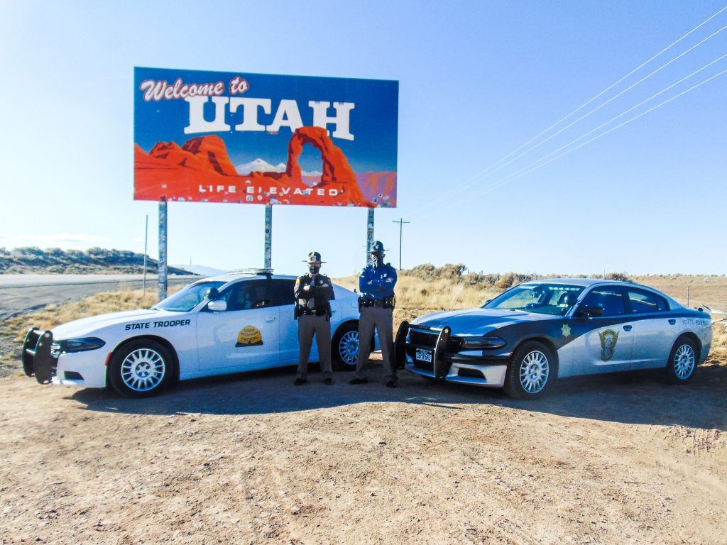 Image shows a UHP vehicle and an CSP vehicle parked in front of the welcome to Utah sign with a UHP Trooper and CO state Trooper standing in front of them.