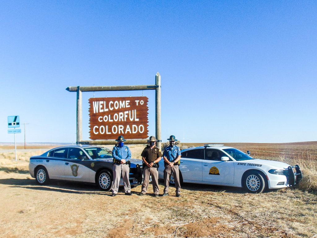 Image shows a UHP vehicle and an CSP vehicle parked in front of the welcome to Colorado sign with a UHP Trooper and CO state Trooper standing in front of them.