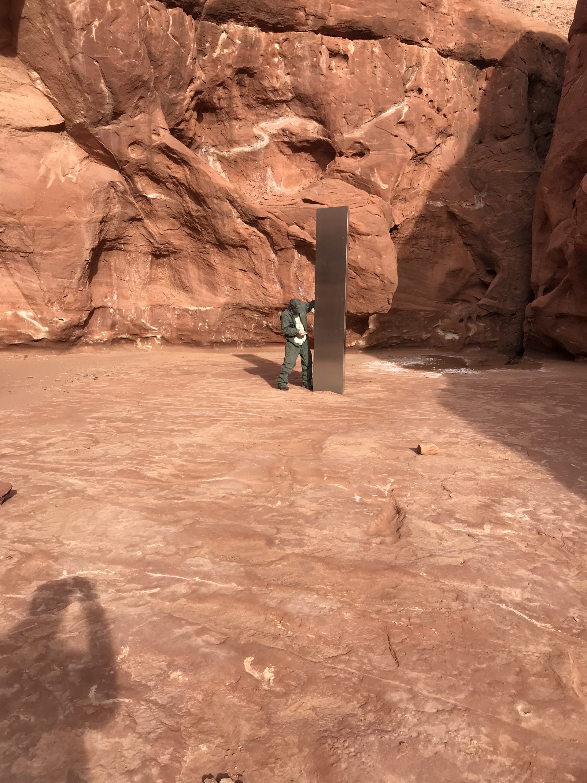 A crew member from the DPS Aero Bureau mission stands next to the metal monolith implanted in the ground in red rock.
