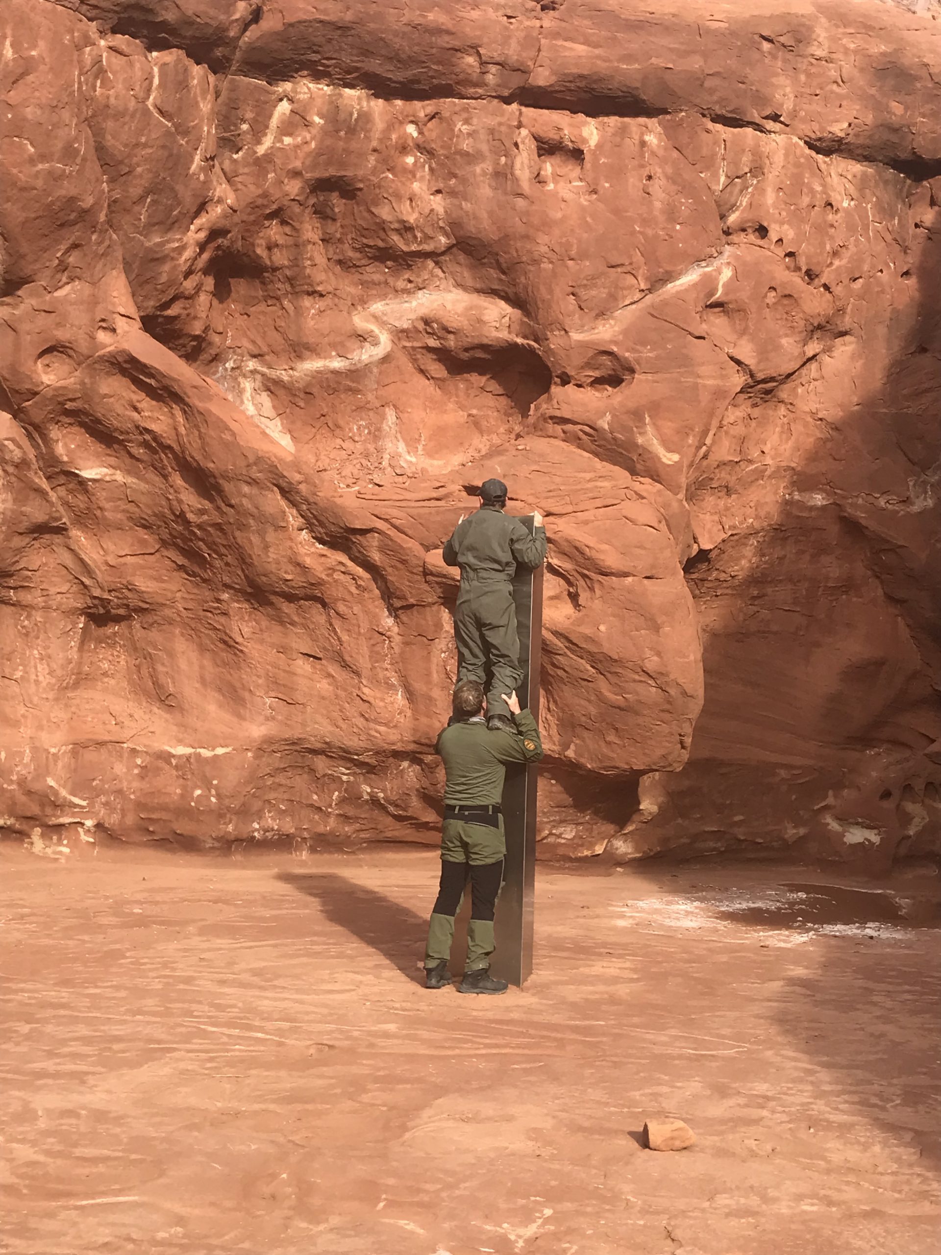 Two crew members - one standing on the other's shoulders - from the DPS Aero Bureau mission stands next to the metal monolith implanted in the ground in red rock.