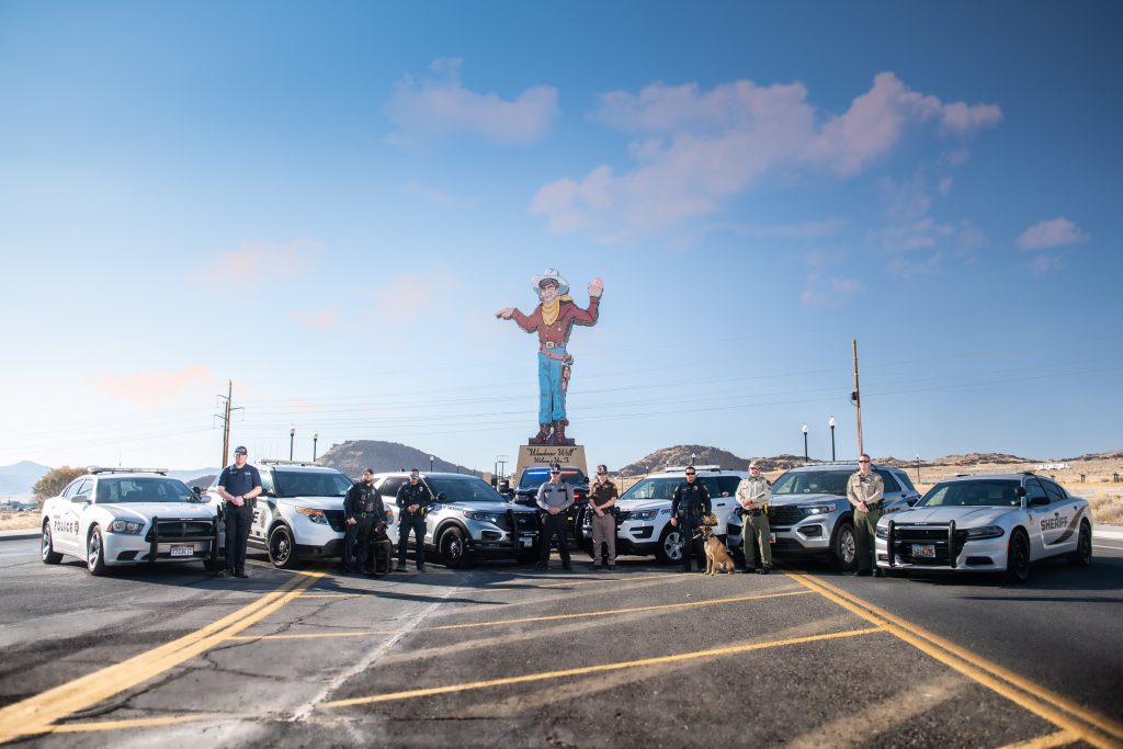 Image shows officers from Tooele County Sheriff's Office, Elko County Sheriff's Office, West Wendover PD, Wendover PD, UHP, NHP standing in front of their vehicles which are positioned in front of the Wendover Will sign.