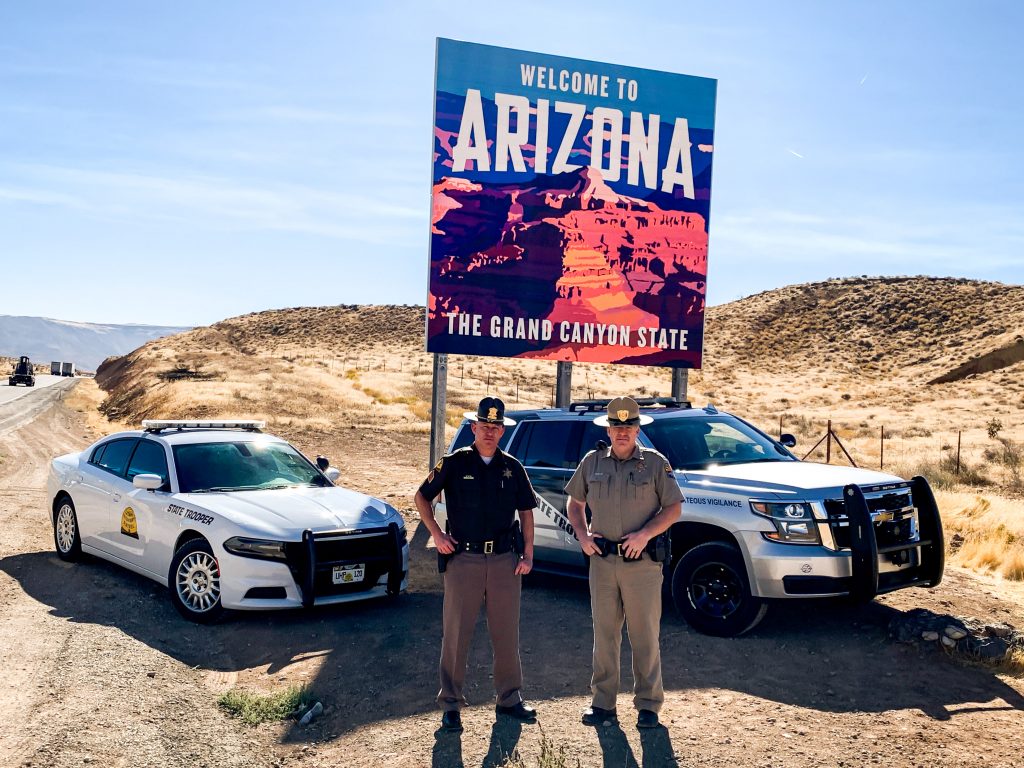 Image shows a UHP vehicle and an AZHP vehicle parked in front of the welcome to Arizona sign with a UHP Trooper and AZ state Trooper standing in front of them.
