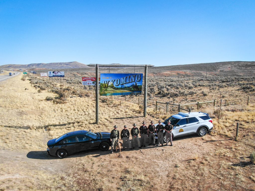 Image shows a group of Wyoming Highway Patrol Troopers and Utah Highway Patrol Troopers standing in front of their vehicles which are positioned in front of the Welcome to Wyoming sign.